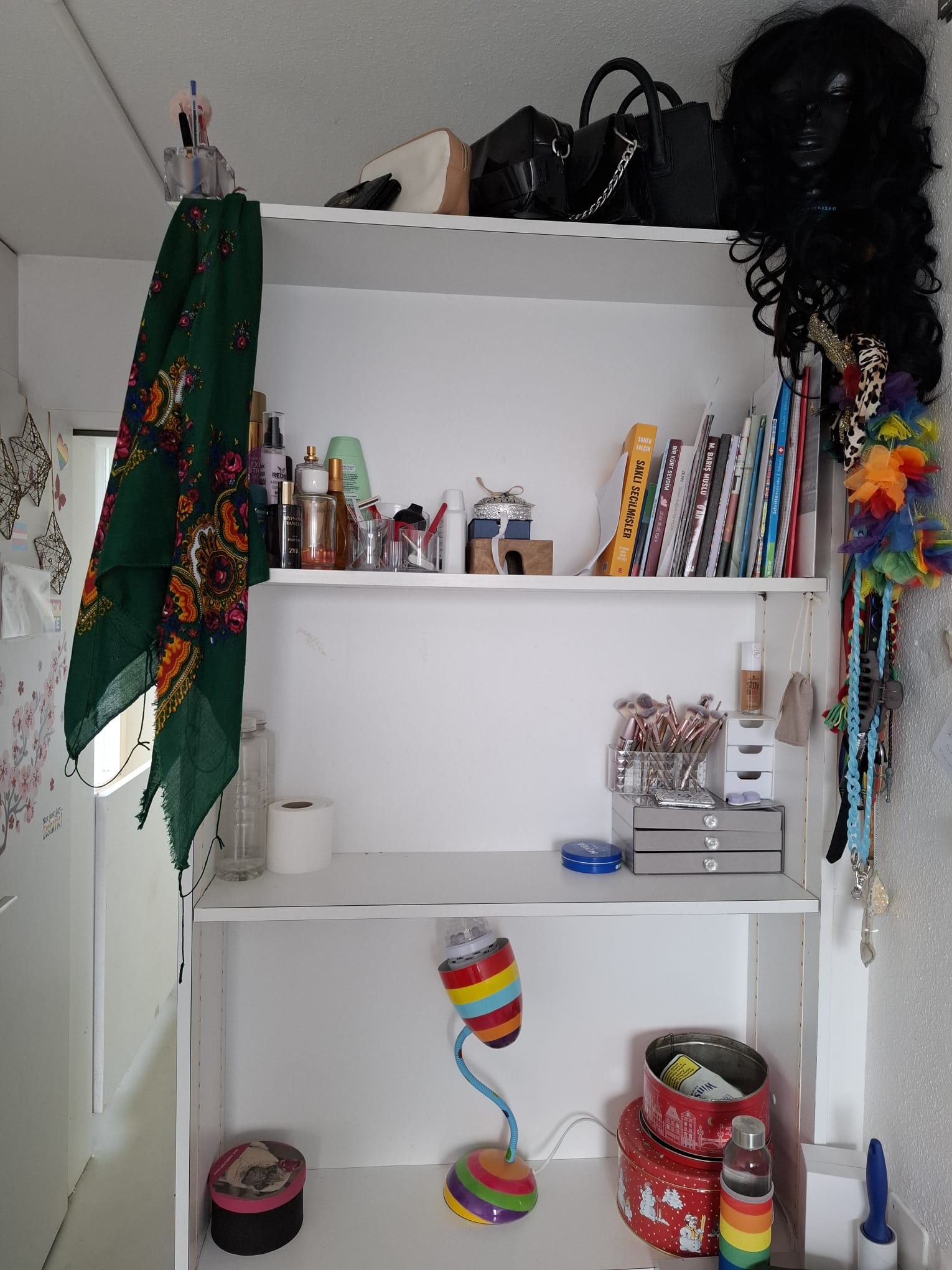 The shelf has three tiers, and each one carries its own quiet story. At the very top are my bags, and right beside them, a wig—its presence quietly waiting. In the corner, a pencil holder, filled with pens ready to write my story. On the shelf beneath the bags are my perfumes. Each bottle holds a whisper from the past, a hidden memory. Even a single drop says, “I am still here.” Across from the perfumes are my books: the ones I’ve read and those connected to Kurdistan. Getting lost among their pages is like taking a breath in my silent world. The books hear the words I cannot tell anyone, they heal my wounds. One tier down, my makeup and brushes. On the side, my jewelry—small but radiant with confidence. Every glance in the mirror creates moments where I can say, “Yes, this is me.” On the bottom tier, a night lamp in the colors of the LGBT flag. Next to it, a small earring box and a bottle of LGBT essence—quiet but bold reminders of my identity. Its light, its color, the presence of the water… they all tell me, “You are here, you are free.” Amid it all, a silence remains; it neither shouts nor is completely still. It simply whispers: “Trust sometimes does not grow in others, but in the small world you have built for yourself."